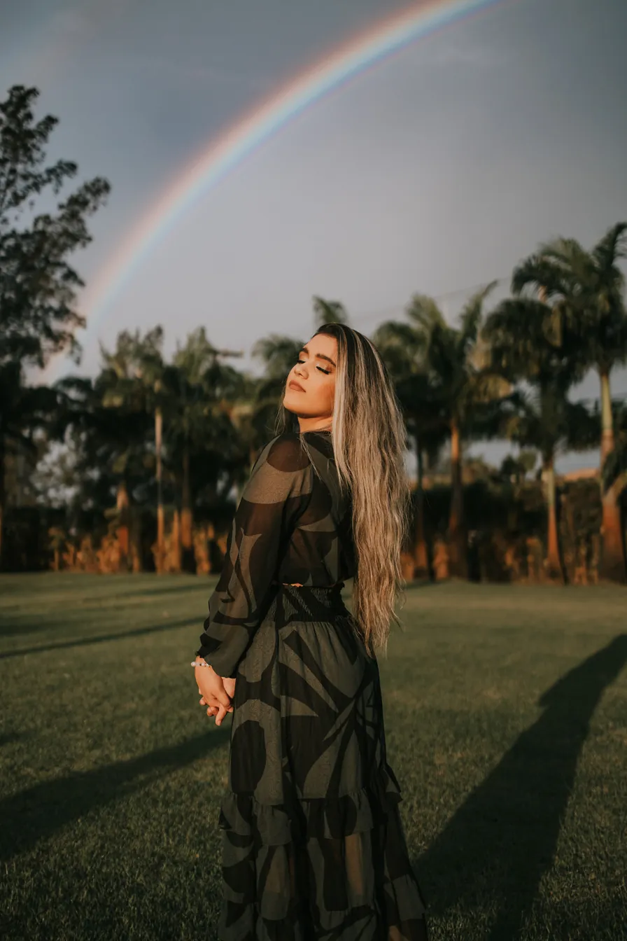 Jovem de vestido escuro num gramado com arco-íris ao fundo